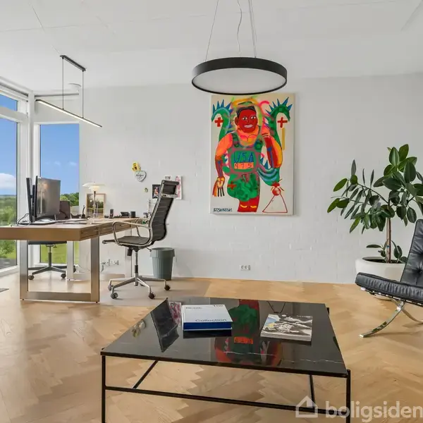 Kontor med skrivebord og stol ved et stort vindue. Et farverigt kunstværk hænger på væggen, en sort lænestol står ved siden af en plante. En sort sofabord står foran.