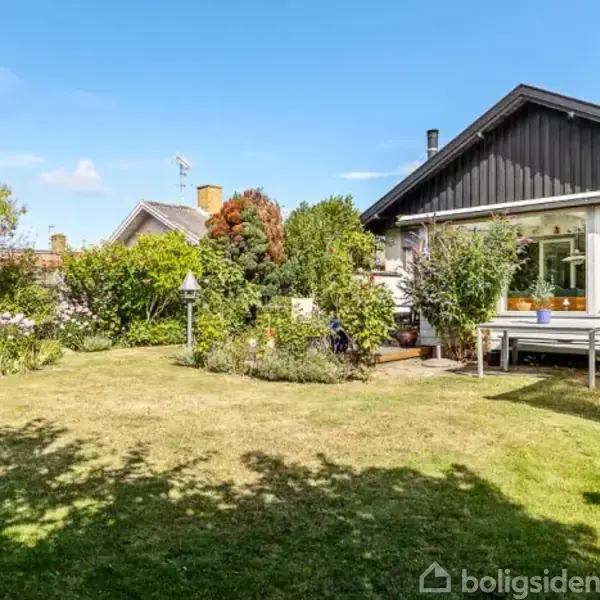 Hus med sort tag og store vinduer vender ud mod en velplejet have med græsplæne og buske under en blå himmel.