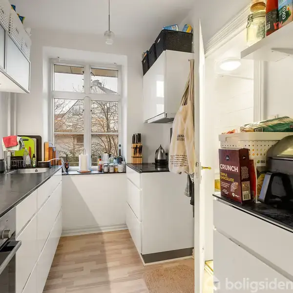 Et smalt køkken har hvide skabe, sorte bordplader, og køkkenredskaber. Der er en vindueskarm med krydderier og rengøringsartikler. En kurv med knækbrød står ovenpå en elkedel.