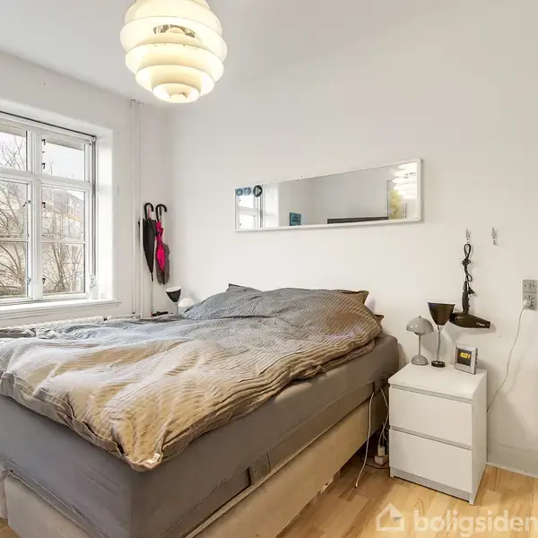 Seng med gråt sengetøj står i et lyst soveværelse. Et vindue til venstre giver udsigt til træer udenfor. Natbord til højre med lampe og dekorationer.