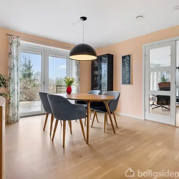 Et spisebord med fire grå stole står i et lyst rum. En stor lampe hænger over bordet. Vinduer og glasdøre giver udsigt til en terrasse og stue.