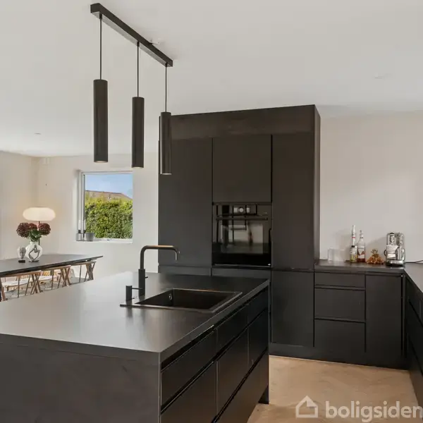 Sort, moderne køkken med ø-køkken og indbygget ovn, vask og armatur. Spisebord med stole ses i baggrunden ved glasdøre, der fører ud til en have med grøn hæk.