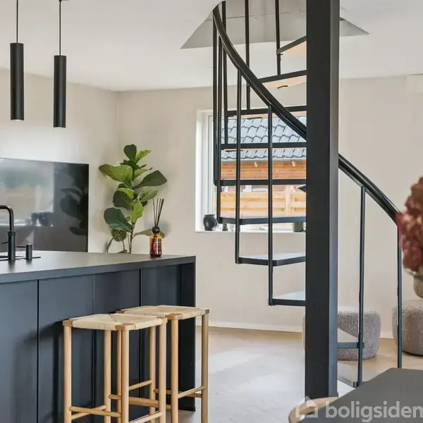 En sort køkkenø med to træstole står i et lyst rum. En vindeltrappe fører til øverste etage, omgivet af minimalistisk indretning og en fladskærm på væggen.