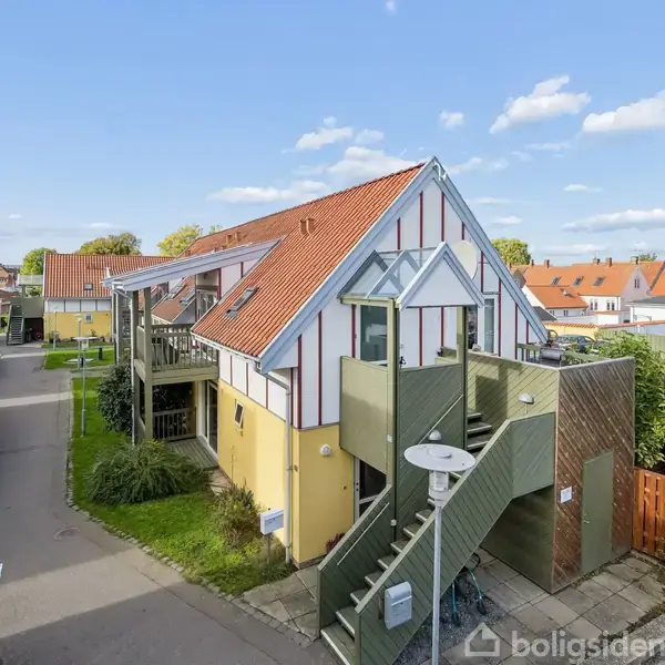Et to-etagers gult hus med rødt tegltag har en grøn trappe op til en balkon. Det ligger i et boligområde med flere lignende huse og en asfalteret vej.
