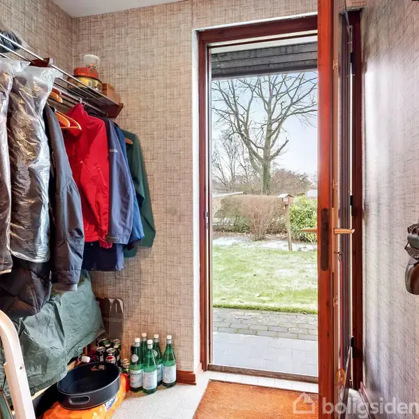 Entré med overtøj hængende; åben dør fører ud til en have med sneplettet græs og træ; flere flasker er placeret på gulvet ved indgangen.