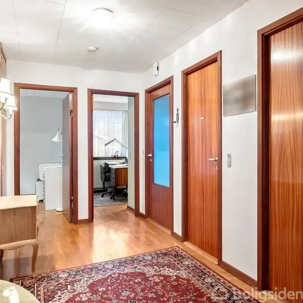 Gang med blanke trægulve, tre lukkede trødøre. Tre åbninger fører til lyse rum. Røde tæppe ligger midt på gulvet. Væggene er dekoreret med lamper og spejl.