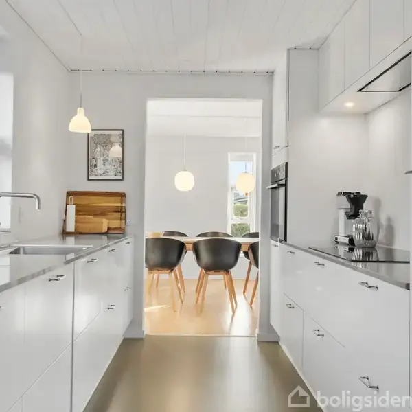 Et lyst, moderne køkken med hvide skabe og indbygget ovn. Rummet har en vask, kaffemaskine og skærebræt. I baggrunden ses en spisestue med stole og et rundt bord.