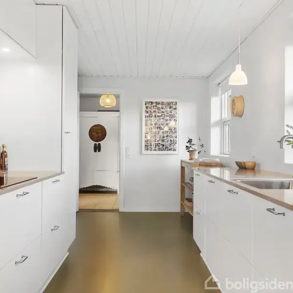 Hvidt moderne køkken med træbordplader, knivblok og planter på bordet. To store vinduer giver naturligt lys. En åben dør fører til en entré med en rund lampe.