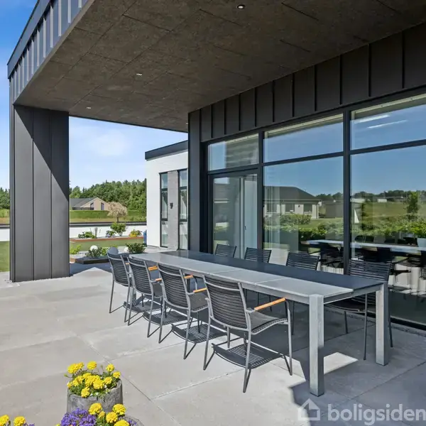 En lang udendørs spisebord står på en terrasse ved et moderne hus. Bordet har otte stole omkring sig, og der er blomster i forgrunden. Terrasse overskygges delvist af tag.