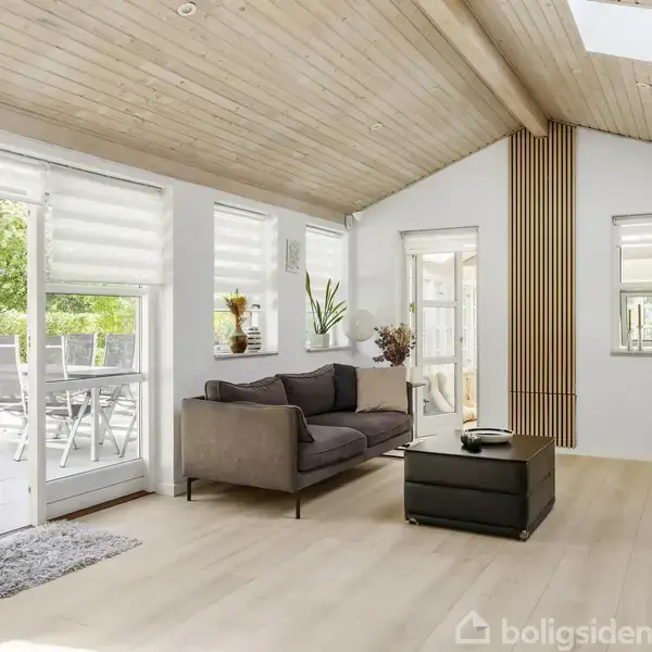 Stue med grå sofa og sort sofabord i moderne nordisk stil. Store vinduer og skydedør åbner til en terrasse med havemøbler. Loftet er skrå og træbeklædt. Jardiner hænger delvist over vinduerne.