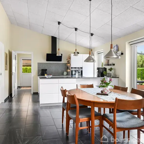 Rundt spisebord med seks stole står i et moderne køkken. Bordet er dækket med en løber og en plante. Store vinduer og en dør giver udsigt til en terrasse med have.