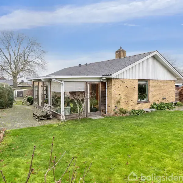 En gulstensbungalow med hvide detaljer står i en frodig have med en stor græsplæne og terrasser. En terrasseoverdækning ses på husets side, og baggrunden har vintertræer.