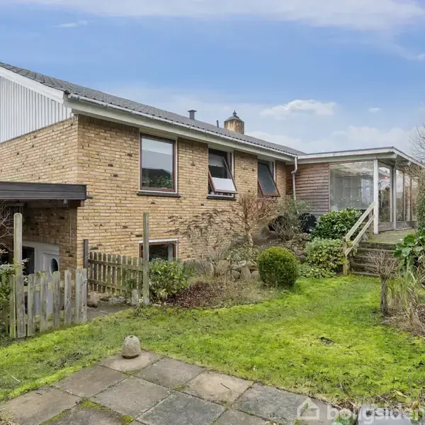 Et enfamiliehus i mursten står stille omgivet af en velplejet have med buske og græsplæne. En stentrappe fører op til en veranda ved siden af huset.