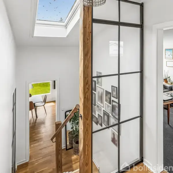 Trappeopgang med trægulv fører til en lys gang, dekoreret med glasvægge og billedrammer. Gennem døre ses stuer med gråt gulvtæppe og moderne møbler. Skylight ovenfor.