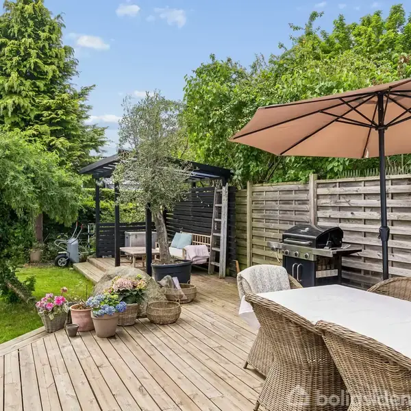 Træterrasse med havemøbler, parasol og grill. Omgivet af blomsterkrukker. I baggrunden ses en havepavillon og grønne træer, med en plæne og cykel ved siden af.
