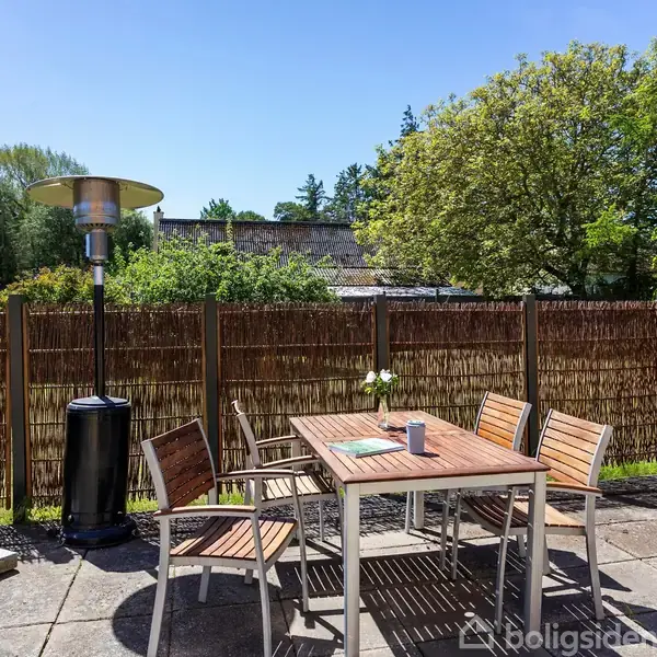 Udendørs havebord med fire stole står på en flisebelagt terrasse. En terrassevarmer står ved siden af, omgivet af træer og et hegn i en solrig have.