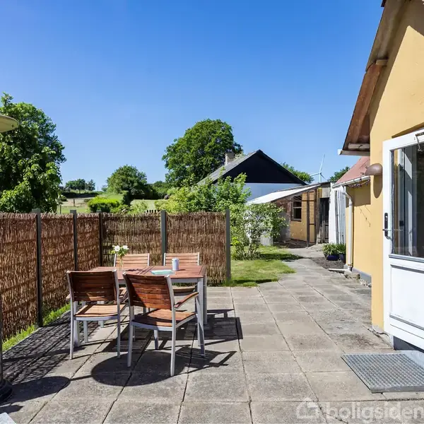 En terrasse med et bord og fire stole står ved siden af et gult hus. En terrassevarmer er placeret nær bordet. Der er grønne træer og en blå himmel i baggrunden.