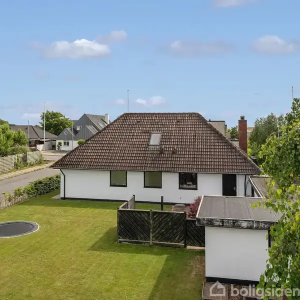 Et hvidt hus med brun tagsten står i en grøn have. En trampolin ses på græsplænen. Beliggende i et villakvarter med veje og flere huse omkring.
