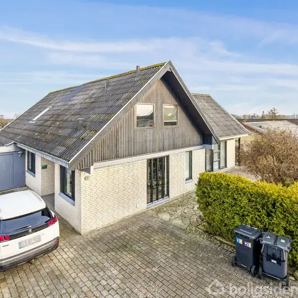Hus står stille på en flisebelagt indkørsel med en hvid bil parkeret til venstre. Omkring er der grønne buske og en blå garage. To skraldespande står ved siden af indkørslen.