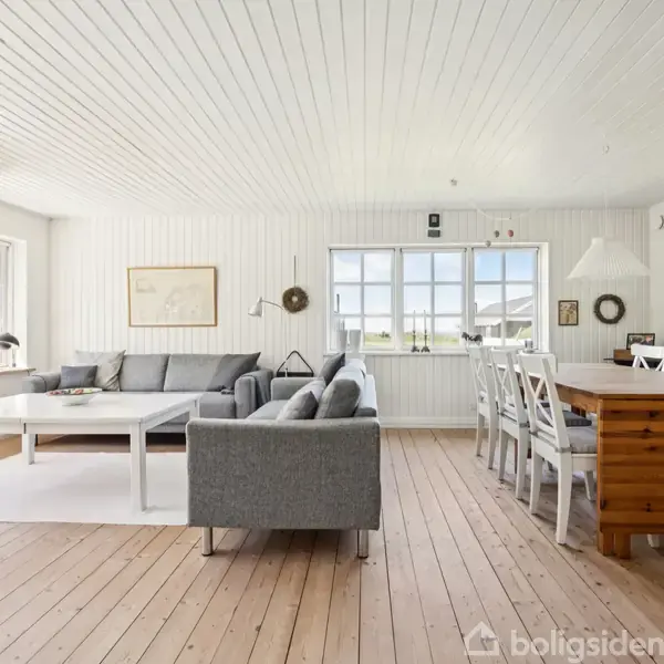 Stue med grå sofa og hvidt sofabord til venstre. Spisebord i træ med hvide stole til højre. Trægulv og hvide lofter skaber en lys atmosfære.