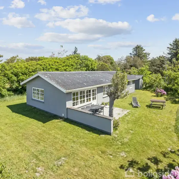 Et gråt enfamiliehus med store vinduer står på en grøn græsplæne. Huset har en terrasse med havemøbler og er omgivet af buske og træer under en blå himmel med spredte skyer.