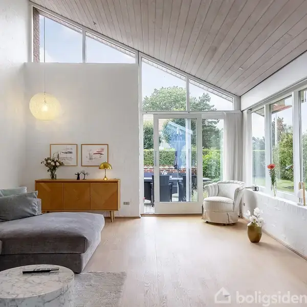 Stue med grå sofa og marmorbord, vendt mod en fladskærms-tv. Store vinduer giver udsigt til en terrasse og have. Moderne indretning med lys trævæg og planter.