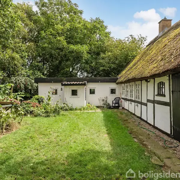 Bindingsværkshus med stråtag står stille i en have med grøn græsplæne. Til venstre er der blomsterbede, og træer omkranser omgivelserne.