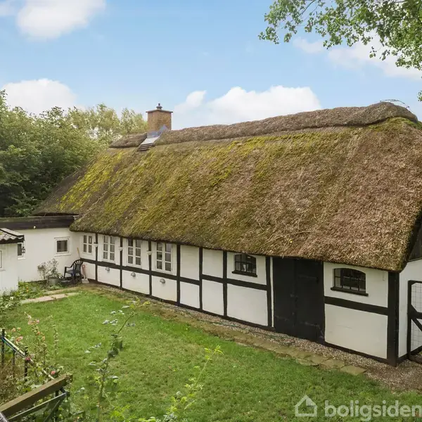 Stråtækt bindingsværkshus med hvid facade og sorte bjælker, står i en grøn have omgivet af træer under en blå himmel med spredte skyer. Ingen synlig tekst.