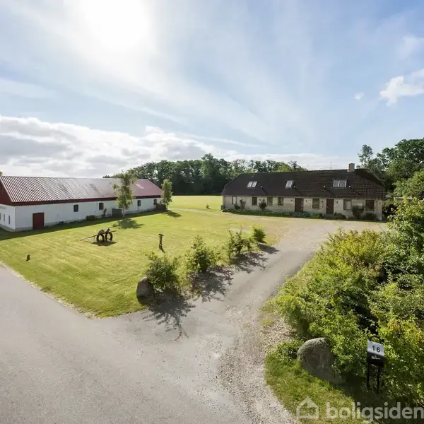 En gård med to bygninger står på en velplejet græsplæne. Den ene bygning er hvid med rødt tag, og den anden er en stor hvid villa. Omgivet af grønne områder og træer.