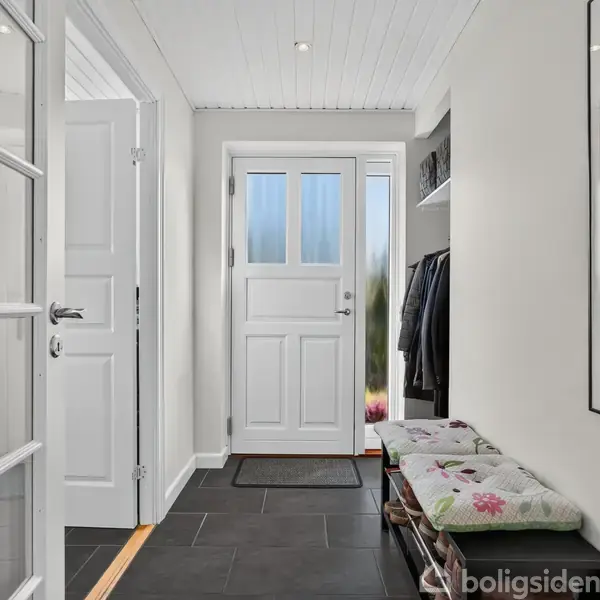 En lys entré med hvide vægge og dør, sorte fliser, og et blomstret sæde på en bænk ved siden af en knagerække med hængende jakker. En plakat hænger på væggen.