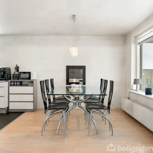 Et glasbord med seks sorte stole står i en spisestue. Køkkenapparater ses i baggrunden, mens store vinduer til højre giver naturligt lys. En lampe hænger over bordet.