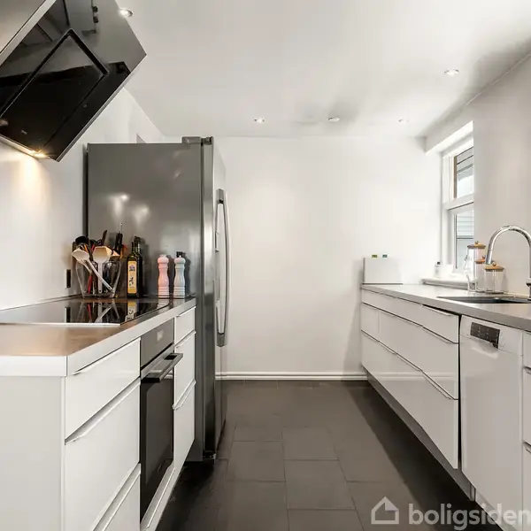 Et moderne køkken med hvide skabe og en stor grå køleskab. Kaffemaskine og køkkenredskaber står på bordpladen. Rummet har sorte fliser og er veloplyst af ovenlys.