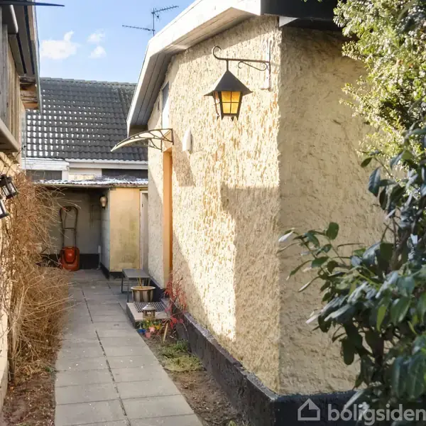 Gangsti fører mellem to bygninger med gule vægge. En sort lanterne hænger på den nærmeste mur. Buske gror tæt op ad den yderste væg.