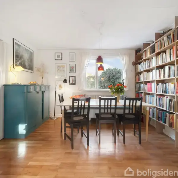 Et spisebord med sorte stole står foran et vindue. En fyldt bogreol står til højre, mens en grøn skænk med billeder hænger til venstre. Sollys oplyser rummet.