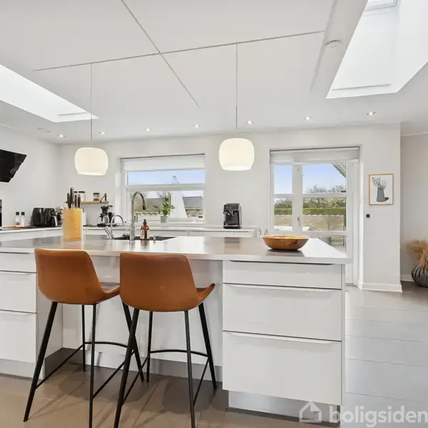 Et moderne køkken med hvid køkkenø, barstole og pendellamper; store vinduer giver naturligt lys, mens et tilstødende rum kan ses i baggrunden.