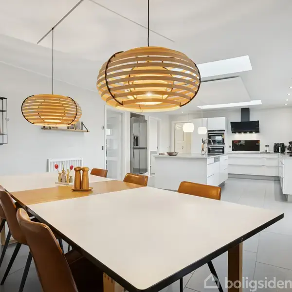 Et spisebord står i midten med stole omkring, belyst af moderne lampeskærme. I baggrunden ses et lyst køkken med hvide skabe og apparater, hvilket skaber en moderne atmosfære.