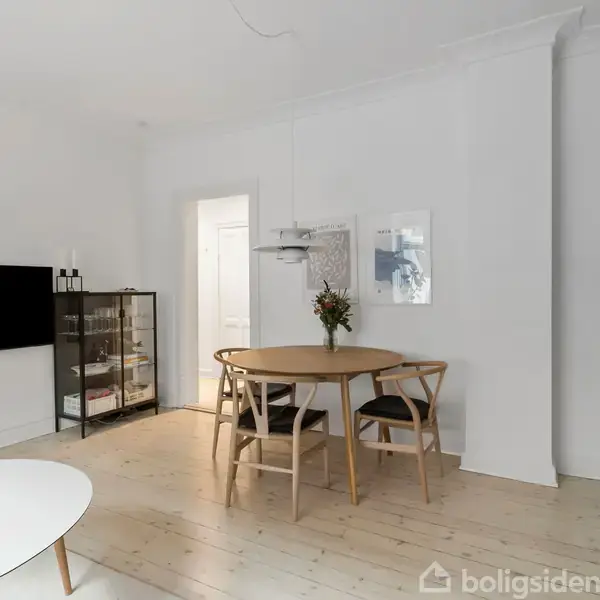 Rundt spisebord med fire stole midt i et lyst rum. Bordet har en vase med blomster. Højttaler hænger på væggen bagved. Et skab står til venstre, køkken til højre.