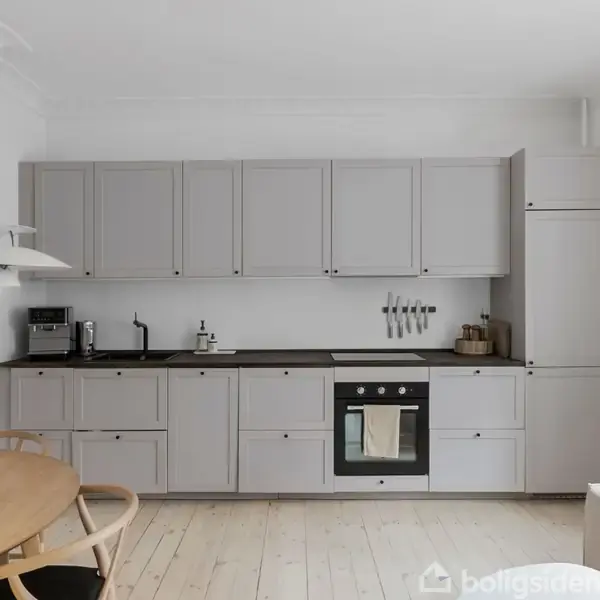 Et lyst, minimalistisk køkken med grå skabe og sorte bordplader. En indbygget ovn og kogetop er midt i skabsvæggen. Ved siden af står et rundt spisebord.