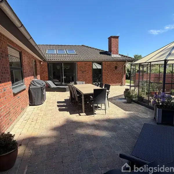 Terrasse med lang spisebord og stole, omgivet af røde murstensvægge. En grill står ved siden af bordet, og en pavillon med planter er placeret i hjørnet. Klar blå himmel.