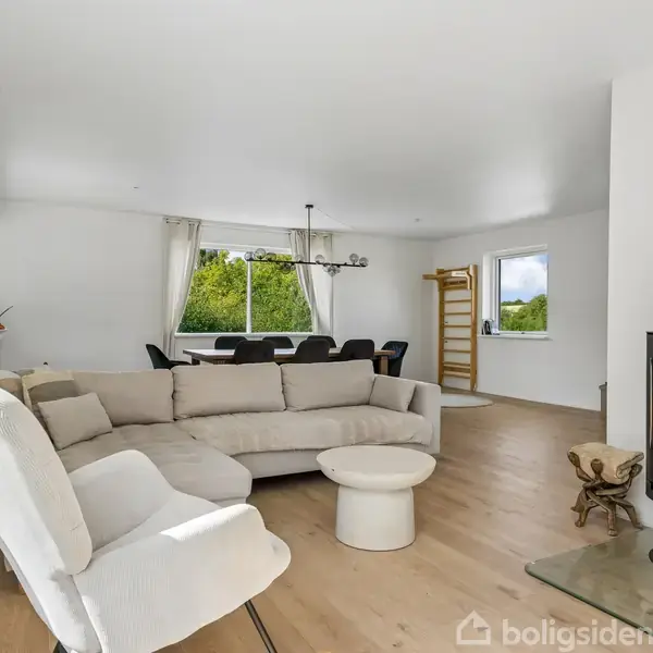 Stue med en hvid lænestol og en beige sofa omkring et rundt bord. Der er en moderne brændeovn til højre, med store vinduer, lysindfald og træer udenfor.