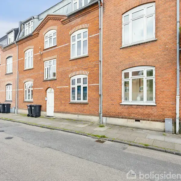 Rød murstensbygning med hvide vinduer i en række. Flere skraldespande står langs fortovet. Bygningen er beliggende på en stille gade med fortov og asfaltvej.