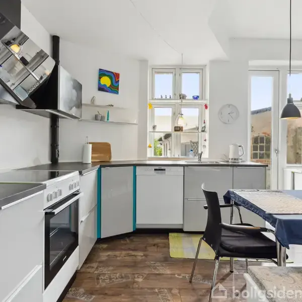 Køkken med hvide skabe og sort komfur. Et sort spisebord med to stole står ved vinduet, hvor der hænger en lampe. Lys trægulv og lys strømmer ind fra vinduerne.