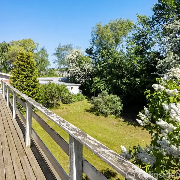 En lang træterrasse strækker sig langs en husfacade med udsigt over en have fyldt med grønne træer og buske under en klar blå himmel.