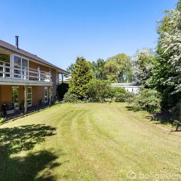 To-etagers hus står stille ved siden af en velholdt græsplæne, omgivet af frodige træer og buske under en klar blå himmel.