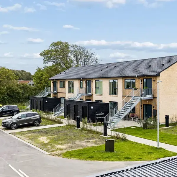 Rækkehus i to etager med udvendige trapper, omgivet af græsarealer. Flere biler parkeret på vejen foran, med træer og en blå himmel i baggrunden.