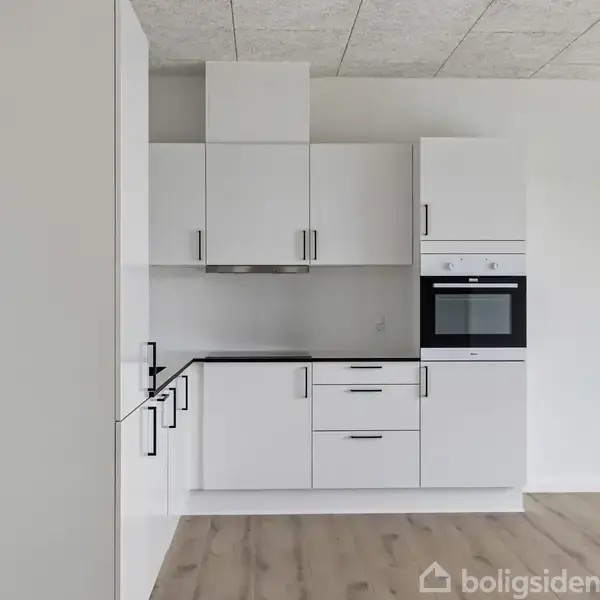 Hvidt moderne køkken med skabs- og skuffemoduler. Ovnen er integreret i et højskab. Placeret i et rum med trægulv og hvide vægge, neutrale nuancer.
