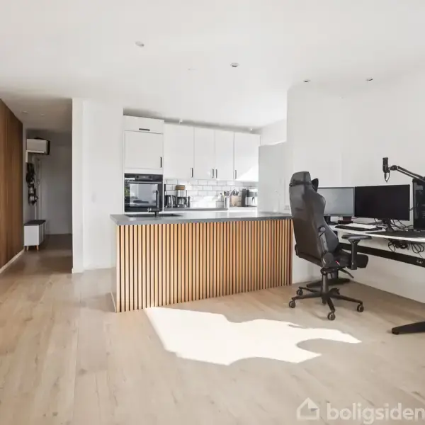Et moderne rum med en køkkenø og en arbejdsstation. Køkkenet har hvide skabe og integrerede apparater. Et skrivebord med en skærm og stol står ved siden af. Gulvet er lyst træ.