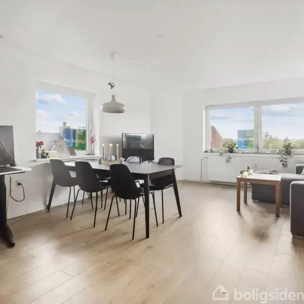 Stue med spisebord og fire sorte stole. En computer står på et skrivebord i venstre hjørne. Området er lyst og moderne med store vinduer.