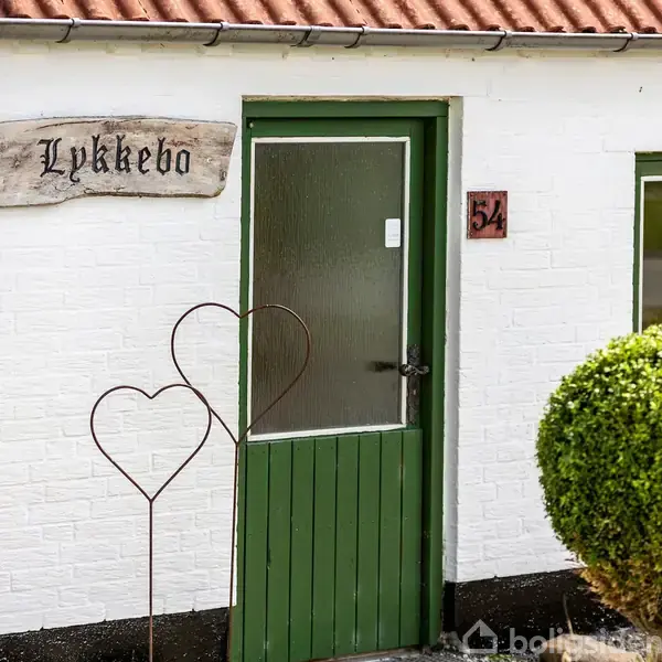 En hvid bygning med grøn dør og vinduer. Et skilt med teksten "Lykkebo" er over døren. To metal hjertefigurer står udenfor. Huset er nummereret 54.