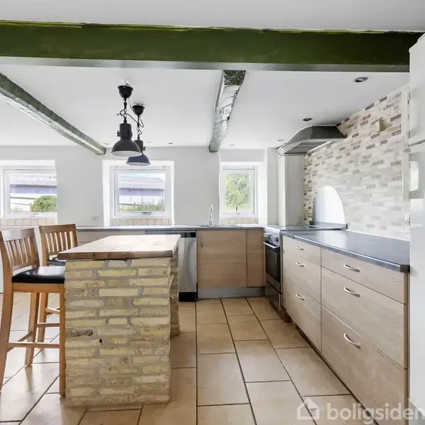 Køkken med træloftsbjælker, et murstensbord med barstole og sort-grå bordplade. Hvide skabe, komfur og opvaskemaskine bagved. Tre vinduer lader lys ind i rummet.